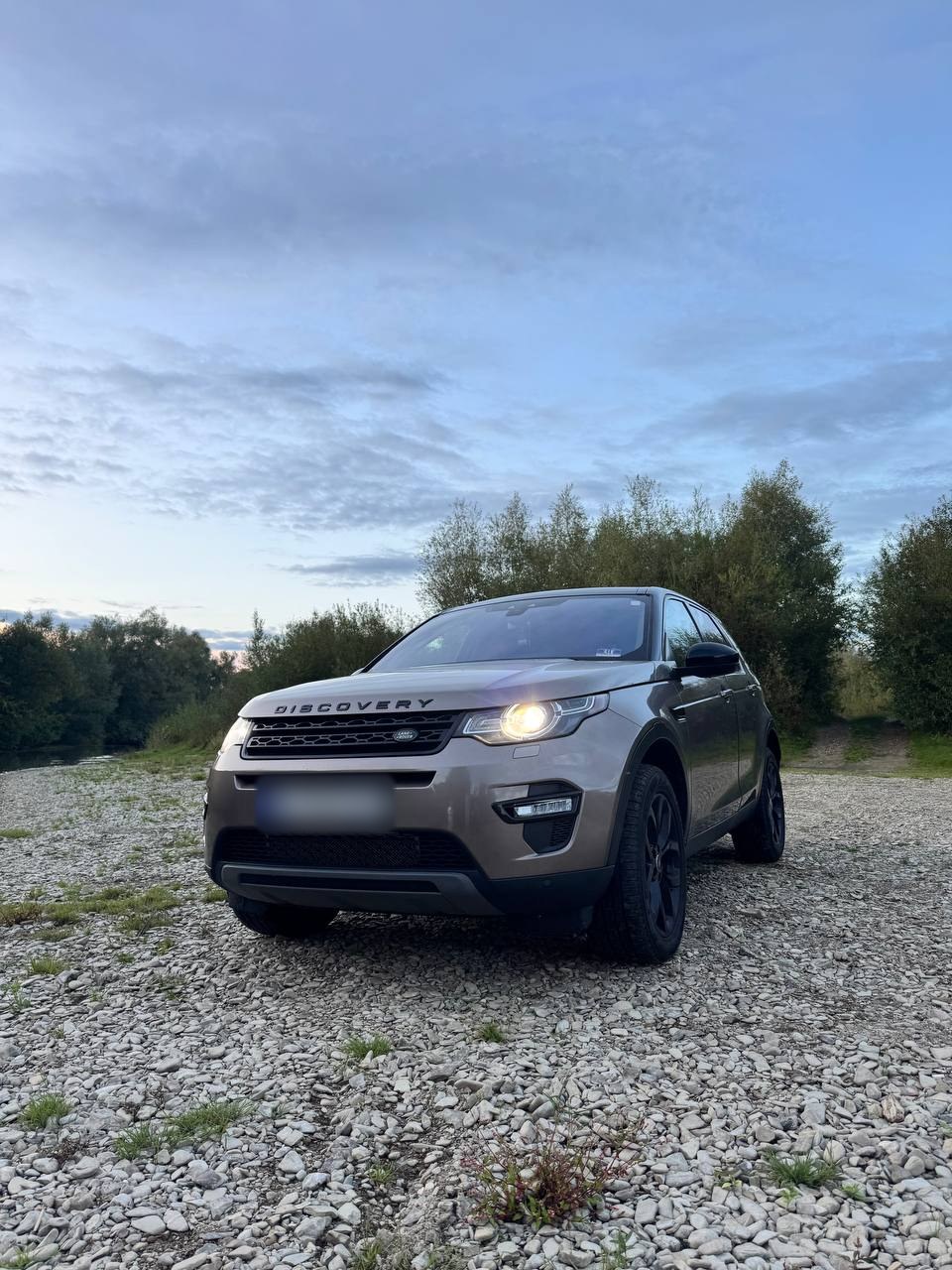 Коричневий Land Rover Discovery Sport учасника клубу з увімкненими фарами на гальці біля річки